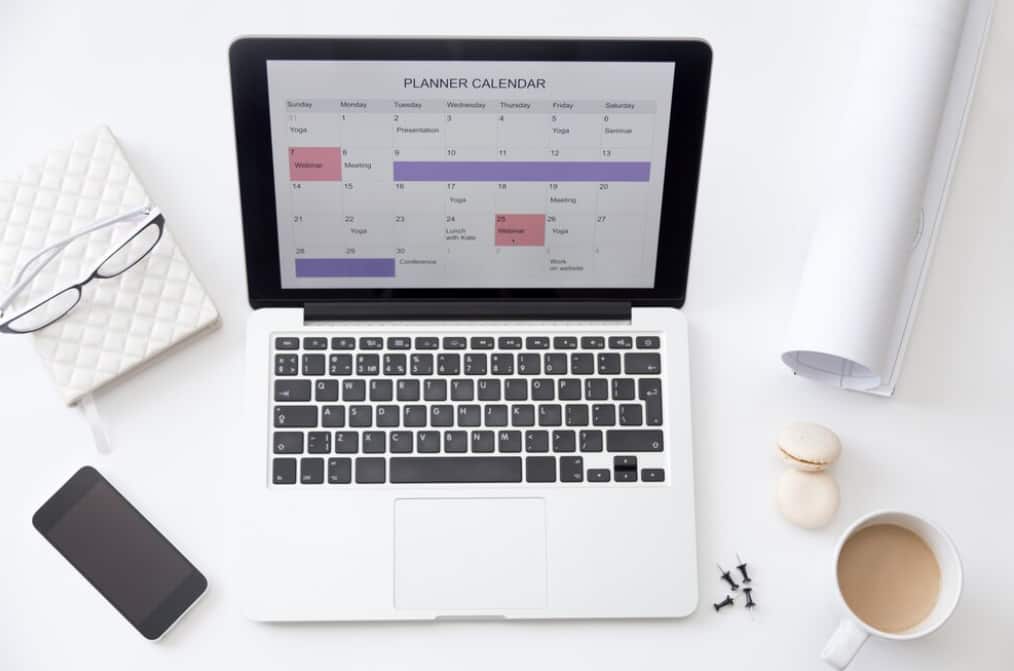 A laptop displaying a weekly planner calendar, with a phone and coffee beside it